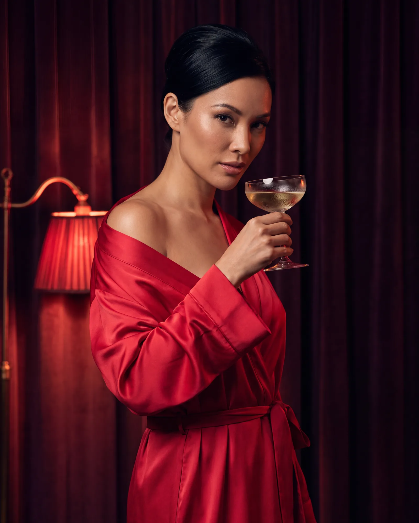 A woman in a red satin robe holding a champagne coupe in front of velvet drapes
